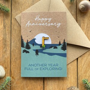 May include: A Happy Anniversary card with a nature-themed design. The card features a landscape with trees, a river, and a sun. The text reads "Happy Anniversary" and "Another year full of exploring!" A pin with a footpath sign is attached.