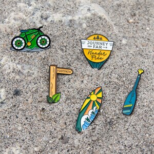 May include: A collection of enamel pins featuring outdoor adventure themes. Designs include a green bicycle, a signpost with "FOOTPATH", a surfboard with a sun and waves, a paddle, and a pin with the text "JOURNEY FAR Wander Free".