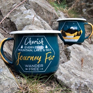 May include: Two teal enamel mugs with white rims and gold handles rest on a rocky surface. One mug features the text "Cherish Every Tree, Mountain, Lake & Sea Journey Far Wander Free." The other mug has a mountain and forest design.