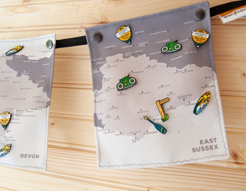 May include: Gray fabric map banner with pins. The banner features a map of East Sussex and Devon, with pins shaped like bicycles, surfboards, and signs. The banner is attached to a wooden background with black ribbon.