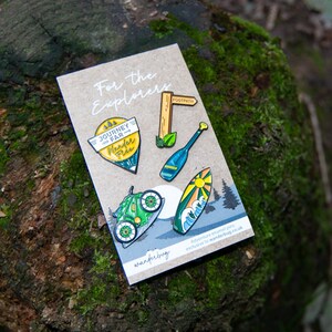 May include: A collection of enamel pins on a cardboard backing, featuring outdoor adventure themes. Designs include a bicycle, surfboard, oar, and a signpost that reads "Footpath". The pins are set against a backdrop of a moss-covered log.