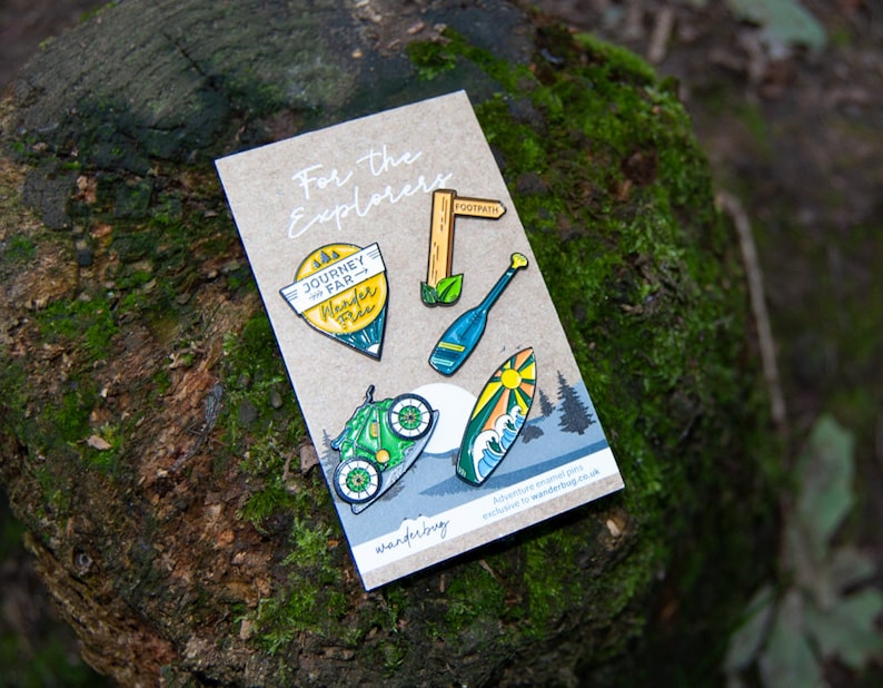 May include: A collection of enamel pins on a cardboard backing, featuring outdoor adventure themes. Designs include a bicycle, surfboard, oar, and a signpost that reads "Footpath". The pins are set against a backdrop of a moss-covered log.
