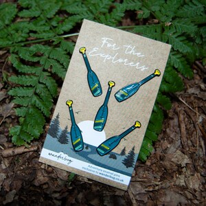 May include: A collection of enamel pins on a kraft paper card. The pins feature a teal paddle design with yellow accents. The card reads "For the Explorers" and includes a small illustration of a forest scene. The background is a bed of green ferns.
