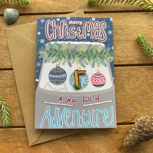 May include: A Christmas card with a car theme, featuring the text "Merry Christmas" and "A year full of Adventure!". The card has a blue background with snowflakes and three hanging ornaments.