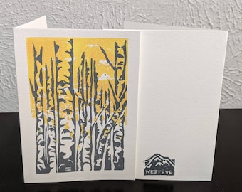 May include: A white folded greeting card featuring a block print illustration of grey birch trees against a bright yellow background. The back of the card displays a small grey mountain graphic above the text WESTEVE in a block print style.