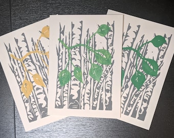 May include: Three rectangular art prints on off-white paper featuring a block-printed design of grey birch tree trunks. Two prints display green leaves on a branch, while the left print features yellow leaves on a branch across the grey trunks.