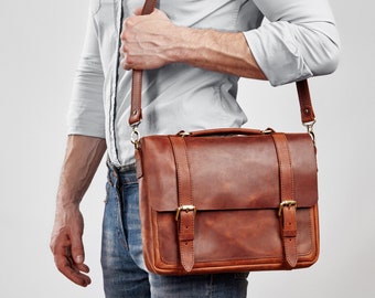 May include: A brown leather satchel with a shoulder strap and a handle. The bag has two buckle closures and a flap. The leather has a rich, warm tone. The bag is carried over the shoulder.