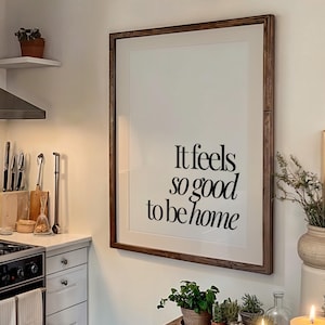 May include: A cozy kitchen scene with a framed print that reads "It feels so good to be home." The room features a stainless steel stove, a wooden table set with plates and a cup, and lit candles, creating a warm, inviting atmosphere.
