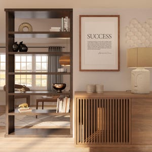 May include: A wooden room divider with five shelves, displaying various decorative objects, including a bowl, books, and a gold animal figurine. The divider is next to a wooden cabinet with a slatted front and a solid wood door. A framed print with the word "SUCCESS" and a quote by Ralph Waldo Emerson is hanging on the wall above the cabinet. A lamp with a beige shade and a face-shaped base sits on top of the cabinet.
