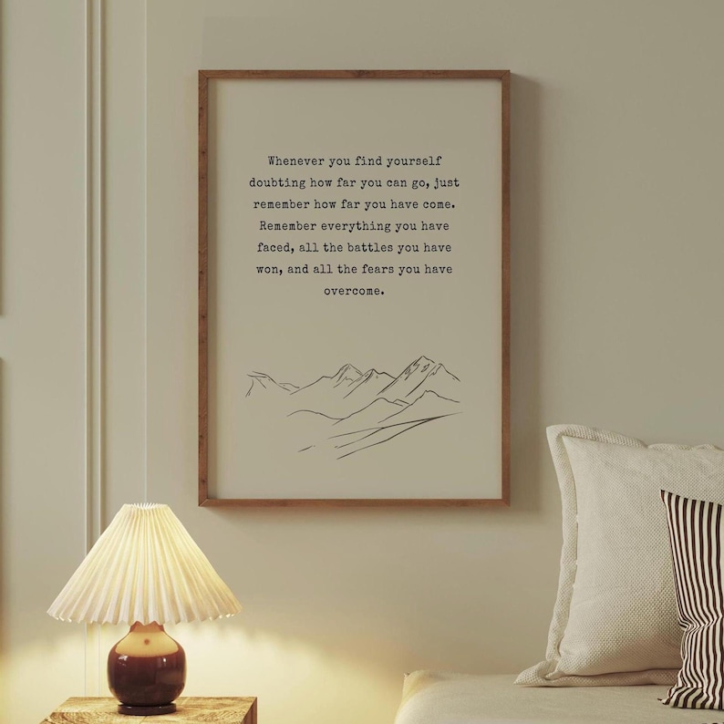 May include: A motivational quote printed on a framed poster with a simple line drawing of mountains at the bottom. The quote reads: "Whenever you find yourself doubting how far you can go, just remember how far you have come. Remember everything you have faced, all the battles you have won, and all the fears you have overcome."