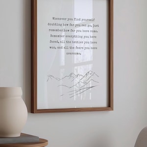 May include: A framed print with a motivational quote about overcoming challenges. The quote reads: "Whenever you find yourself doubting how far you can go, just remember how far you have come. Remember everything you have faced, all the battles you have won, and all the fears you have overcome." The print features a simple line drawing of mountains.