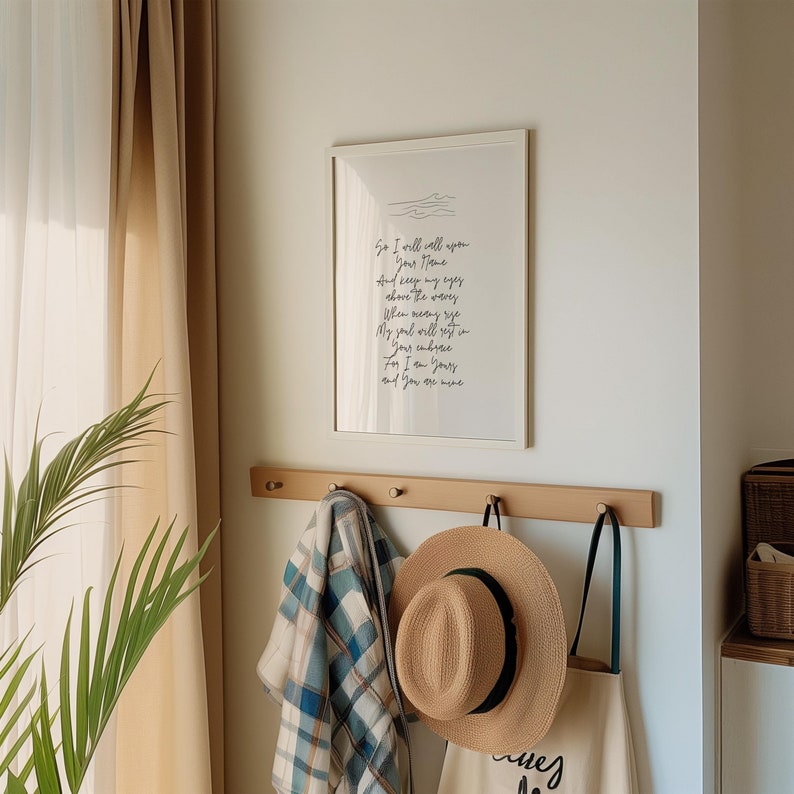 Op de afbeelding: Een wit ingelijst print met een handgeschreven gedicht over liefde en thuis. De print hangt aan een muur met een houten kapstok eronder. De kapstok heeft een geruit deken, een strohoed en een witte draagtas eraan.