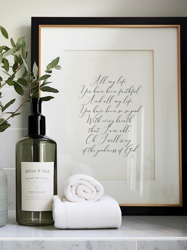 Op de afbeelding: Een ingelijst afdruk met een zwart en gouden frame, met een handgeschreven citaat over de goedheid van God. De afdruk staat op een wit aanrecht met een groene glazen fles en twee witte handdoeken.