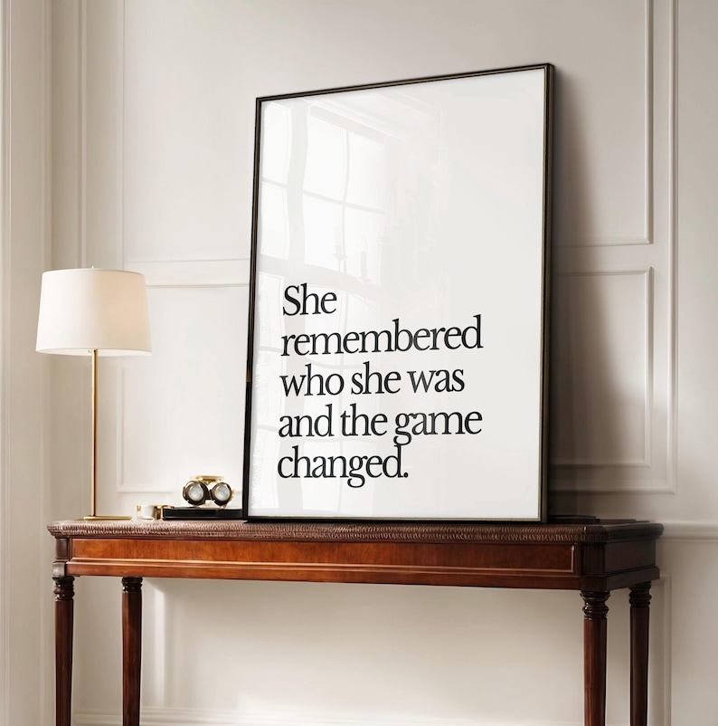 May include: Black and white typography print with the quote "She remembered who she was and the game changed." The print is framed in a black frame and is displayed on a wooden console table.
