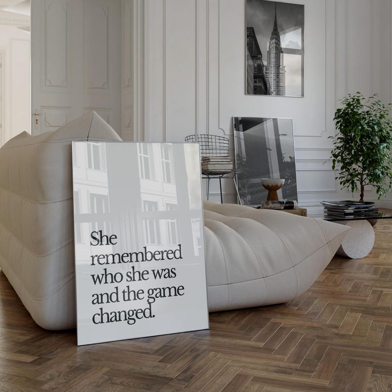 May include: A black and white framed print with the text "She remembered who she was and the game changed." The print is hanging on a white wall in a room with a white couch and a plant.