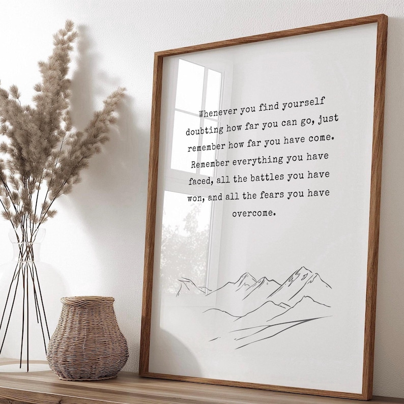 May include: A motivational quote printed on a white background in a wooden frame. The quote reads: "Whenever you find yourself doubting how far you can go, just remember how far you have come. Remember everything you have faced, all the battles you have won, and all the fears you have overcome." A simple line drawing of mountains is at the bottom of the quote.