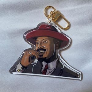 May include: A clear acrylic keychain featuring a smiling man wearing a red hat, suit, and tie. The keychain has a gold-colored clasp and ring. The man is depicted with a mustache and goatee.