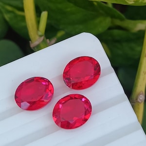 May include: Three oval-shaped, bright red gemstones on a white surface. The gemstones are smooth and shiny.