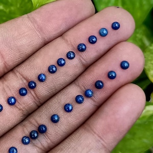 May include: A collection of small, round, deep blue gemstones, possibly sapphires or lapis lazuli, are arranged on a hand. The stones are evenly spaced in rows and columns, showcasing their polished, reflective surfaces. The background is a soft green.