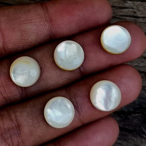 May include: Five round, white mother-of-pearl cabochons with a pearlescent sheen. Each cabochon has a slightly domed surface and a subtle, warm, yellow-toned edge. The cabochons are displayed against a dark, textured background.
