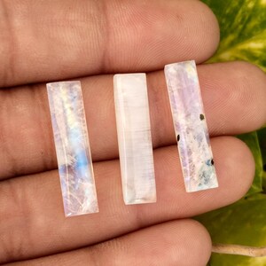 May include: Three rectangular moonstone gemstones with iridescent blue, purple, and white hues. The gemstones are held in a hand, with green foliage in the background. The stones are polished and have a smooth surface.