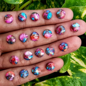 Könnte beinhalten: Nahaufnahme einer Hand, die eine Sammlung runder Edelsteine hält. Die Steine zeigen ein Mosaikmuster in Rosa, Blau und Gold. Die polierten Steine zeigen lebendige Farben und komplizierte Muster. Jeder Edelstein hat einen Durchmesser von etwa 1 cm.