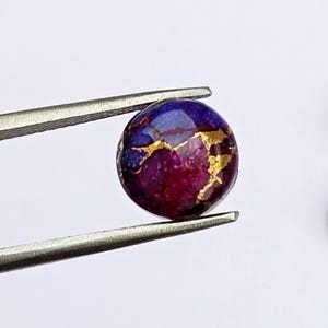 May include: A round gemstone held by tweezers. The stone features a mix of colors, including deep purple, blue, and gold. The gemstone has a polished surface and is likely used for jewelry or decorative purposes.