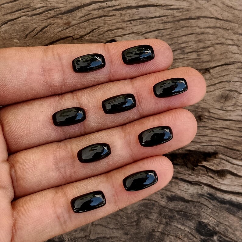May include: A collection of nine polished, rectangular black gemstones. The stones are smooth and reflective, with rounded edges. The gemstones are displayed on a hand, with a wooden surface in the background.