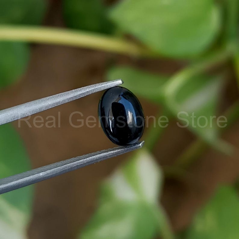 May include: A polished, oval-shaped black gemstone is held by silver tweezers. The gemstone reflects light, showcasing its smooth surface. The text "Real Gemstone Store" is visible in the background.