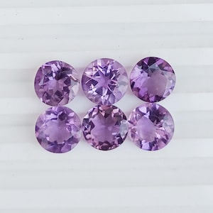 May include: Six round, faceted amethyst gemstones in varying shades of purple. The gemstones are arranged in two rows of three, each with a brilliant cut that reflects light. The gems are set against a white background.