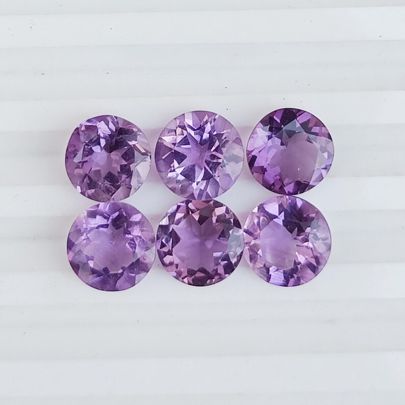 May include: Six round, faceted amethyst gemstones in varying shades of purple. The gemstones are arranged in two rows of three, each with a brilliant cut that reflects light. The gems are set against a white background.