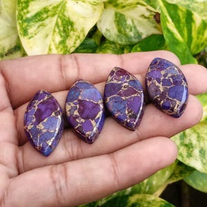 May include: Four polished, teardrop-shaped gemstones with a vibrant mix of purple, blue, and gold hues. The stones are held in a hand, showcasing their unique patterns and textures. The gemstones are ideal for jewelry making.
