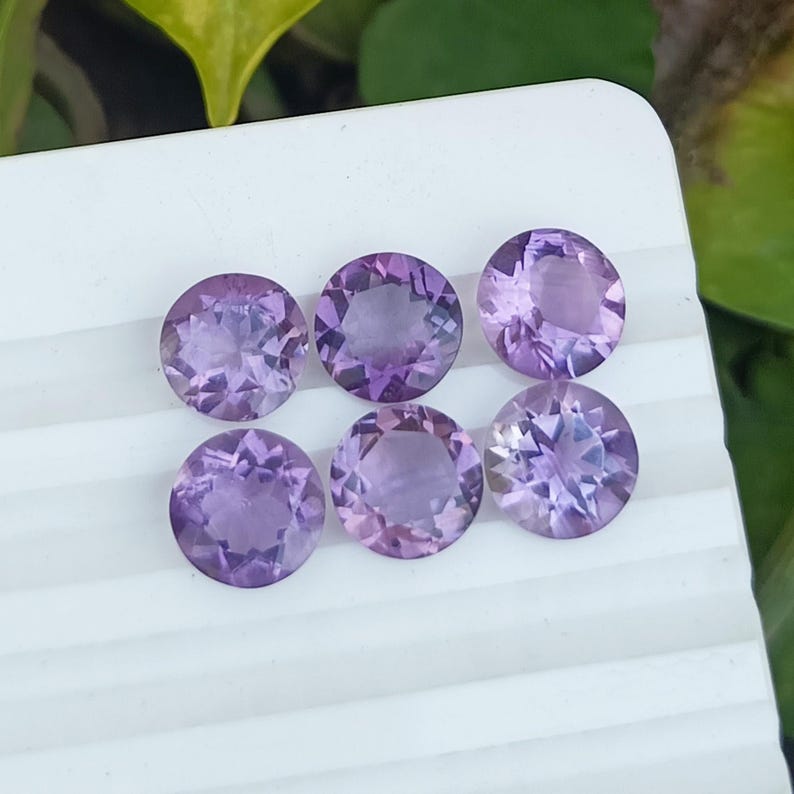 May include: Six round, faceted amethyst gemstones in varying shades of purple and lavender. The gemstones are displayed on a white surface, with a blurred green background.