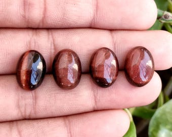 May include: Four oval-shaped, polished gemstones with a reddish-brown hue. The stones have a smooth, reflective surface, and are displayed against a neutral background. The gemstones are likely used for jewelry making or collecting.