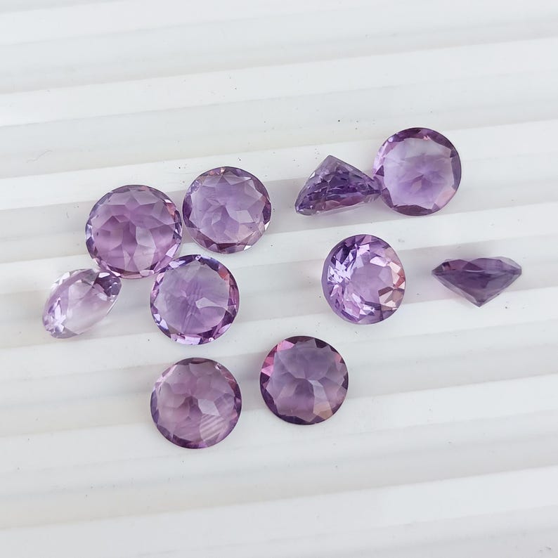May include: A collection of faceted amethyst gemstones in various shapes, including round, oval, and triangular. The gemstones display a range of purple hues, from light lavender to deep violet. The gemstones are arranged on a white surface.