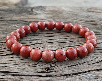 May include: A red jasper beaded bracelet. The bracelet features polished, round beads in varying shades of red and brown. The beads are strung together, forming a complete circle. The bracelet is displayed on a weathered wooden surface.