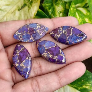 May include: Four oval-shaped gemstones with a mix of purple, blue, and gold colors. The gemstones are held in a hand, and the background is blurred green foliage.