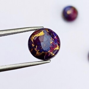 May include: A round, polished gemstone with a deep purple base color, accented with gold veining. The gemstone is held by a pair of silver tweezers against a white background. Additional gemstones are visible in the background.