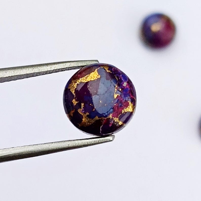 May include: A round, polished gemstone with a deep purple base color, accented with gold veining. The gemstone is held by a pair of silver tweezers against a white background. Additional gemstones are visible in the background.
