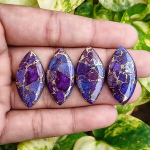 May include: Four marquise-shaped gemstones with a mosaic pattern of deep purple, blue, and gold. The stones are held in a hand, showcasing their polished surfaces and unique color variations. These gemstones are ideal for jewelry making.