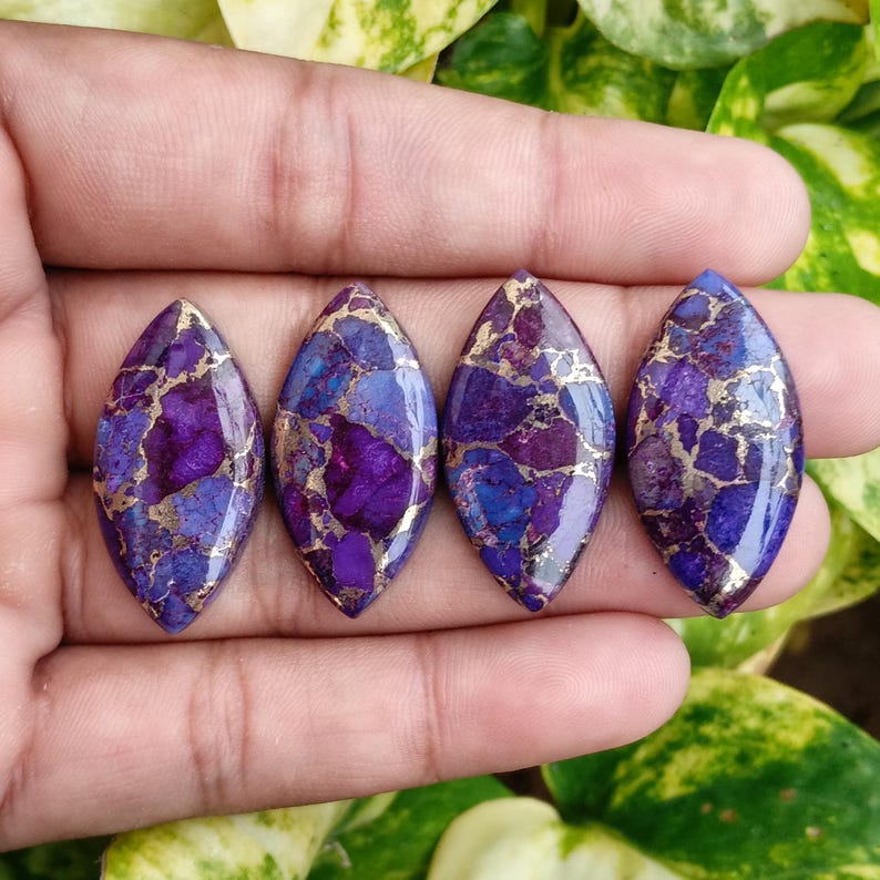 May include: Four marquise-shaped gemstones with a mosaic pattern of deep purple, blue, and gold. The stones are held in a hand, showcasing their polished surfaces and unique color variations. These gemstones are ideal for jewelry making.