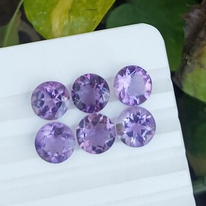 May include: Six round, faceted amethyst gemstones in varying shades of purple are arranged on a white surface. The gemstones are cut with multiple facets, reflecting light and creating a sparkling effect. The gems are a beautiful addition to any jewelry collection.