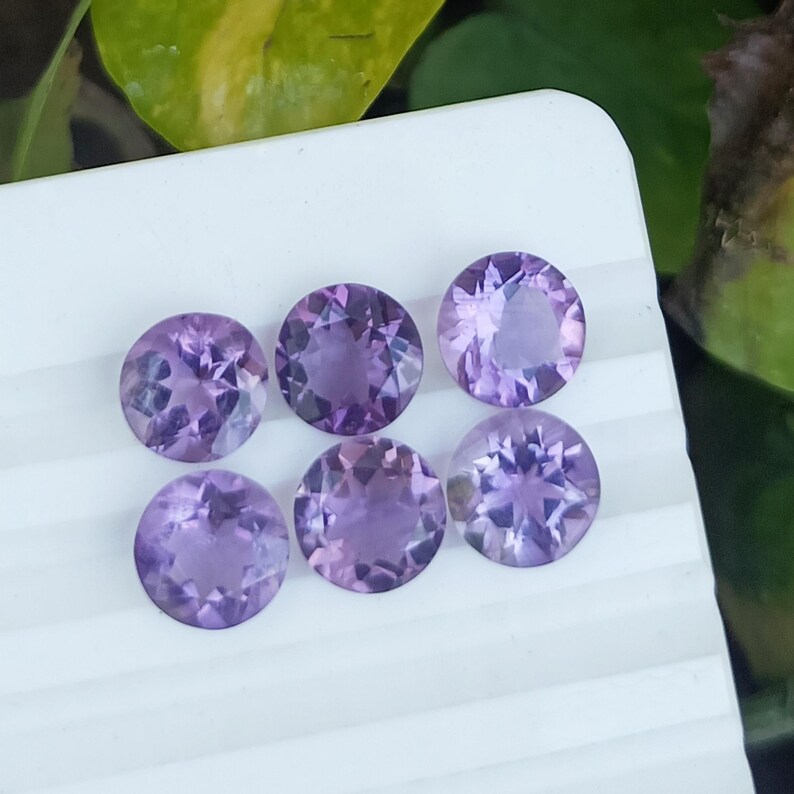 May include: Six round, faceted amethyst gemstones in varying shades of purple are arranged on a white surface. The gemstones are cut with multiple facets, reflecting light and creating a sparkling effect. The gems are a beautiful addition to any jewelry collection.