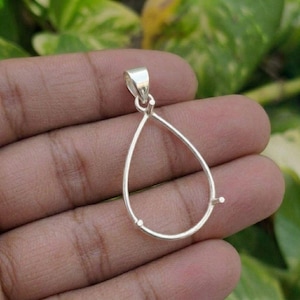 May include: A silver-colored metal pendant setting shaped like a teardrop. The frame is open in the center and features a small bail at the top for attachment to a chain. The setting includes three small prongs at the base and sides for securing a stone.