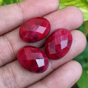 May include: Three faceted, oval-shaped, deep red gemstones. The gems are held in a hand, with a green leafy background. The gems have a polished, reflective surface, and are likely rubies or similar precious stones.