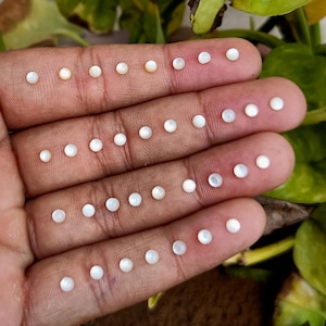 May include: A hand displays several small, round, white pearls. The pearls are arranged in neat rows across the fingers and palm. The pearls have a lustrous, iridescent sheen, reflecting light. The background is blurred, with green foliage visible.