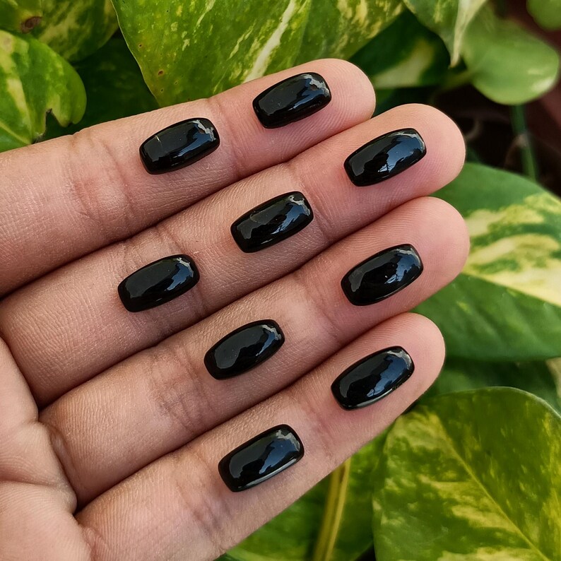 May include: A collection of nine polished, rectangular black gemstones. Each stone has rounded edges and a glossy surface, reflecting light. The gemstones are displayed on a hand, with green and yellow foliage in the background.