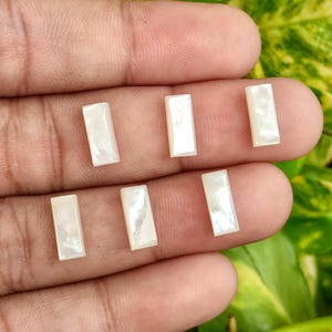 Puede incluir: Siete cabujones rectangulares de nácar iridiscente. Cada cabujón es de color blanco crema con una superficie sutil y brillante. Las piedras preciosas se muestran sobre un fondo color piel, con follaje verde y amarillo en el fondo.