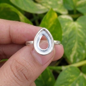 May include: A silver ring featuring a teardrop-shaped bezel setting. The metal band is smooth and polished, leading to a hollow, pear-shaped frame designed to hold a gemstone or inlay. The setting is open, showcasing a clean, metallic finish.