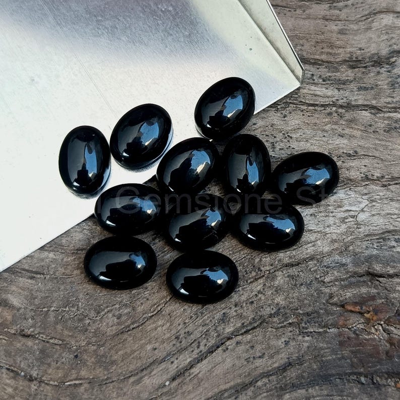 May include: A collection of polished, oval-shaped black gemstones. The stones are arranged on a wooden surface, reflecting light and appearing glossy. The gemstones are a deep, solid black color.